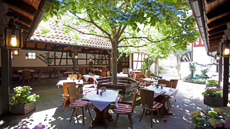 Ein gemütlicher Innenhof mit Tischen und Stühlen unter einem Baum, umgeben von Fachwerkhäusern. Die Tische sind mit gestreiften Tischdecken gedeckt., © Hotel Hirsch
