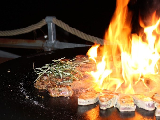 Ein Grill mit brennendem Feuer, Fleischstücken und Rosmarinzweigen im Dunkeln. Im Hintergrund ein Seilgeländer., © Cool-Tours StattReisen Ein Grill mit brennendem Feuer, Fleischstücken und Rosmarinzweigen im Dunkeln. Im Hintergrund ein Seilgeländer., © Cool-Tours StattReisen