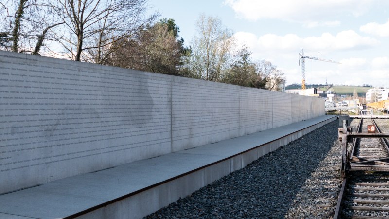 Eine lange Betonwand mit eingravierten Namen steht neben stillgelegten Bahngleisen unter einem bewölkten Himmel., © Foto-Kunst Andreas Keller Eine lange Betonwand mit eingravierten Namen steht neben stillgelegten Bahngleisen unter einem bewölkten Himmel., © Foto-Kunst Andreas Keller