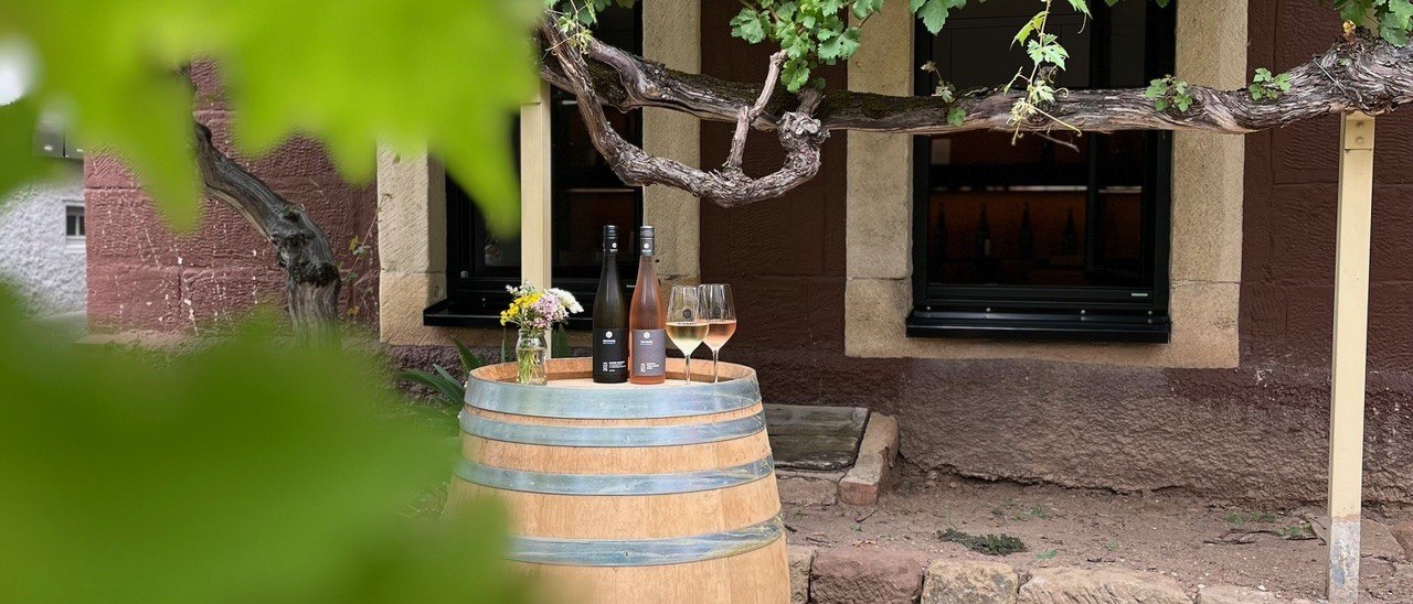Ein Weinfass dient als Tisch für zwei Weinflaschen und Gläser, umgeben von Weinreben vor einem Fenster., © Weingut Schwarz Ein Weinfass dient als Tisch für zwei Weinflaschen und Gläser, umgeben von Weinreben vor einem Fenster., © Weingut Schwarz