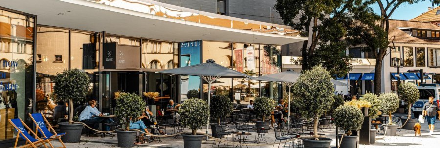 Au&szlig;enbereich eines Caf&eacute;s namens Eduard's mit Tischen, St&uuml;hlen und Sonnenschirmen. Menschen sitzen und entspannen, umgeben von Topfpflanzen., &copy; SMG, Sarah Schmid