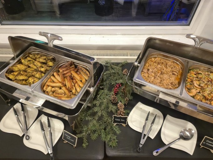 Ein Buffet mit verschiedenen Speisen wie Gem&uuml;se, W&uuml;rstchen und Pulled Pork, dekoriert mit Tannenzweigen und Zapfen, bereit f&uuml;r ein Winter-BBQ., &copy; Cool-Tours StattReisen