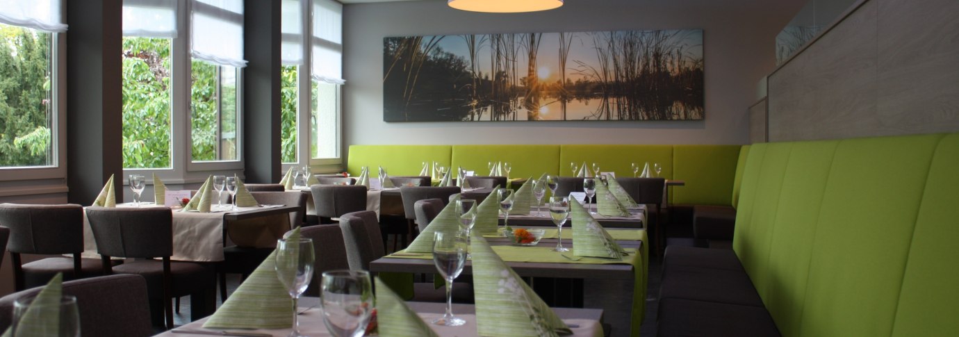 Modernes Restaurant mit gedeckten Tischen, grünen Servietten und großen Fenstern. An der Wand hängt ein Bild mit Sonnenuntergangsmotiv., © Hotel am Schlossberg Herrenberg