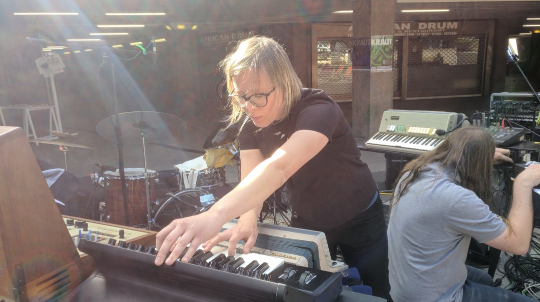 Eine Frau spielt konzentriert auf einem Keyboard bei einem Outdoor-Konzert. Neben ihr arbeitet ein Mann an technischen Ger&auml;ten. Sonnenstrahlen fallen auf die Szene., &copy; privat