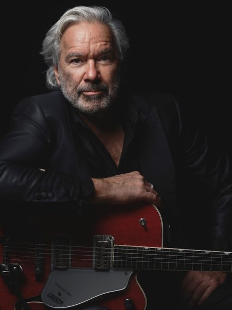 A man with gray hair and beard in a black suit leans on a red electric guitar in front of a black background., © Theaterhaus Stuttgart e.V. A man with gray hair and beard in a black suit leans on a red electric guitar in front of a black background., © Theaterhaus Stuttgart e.V.