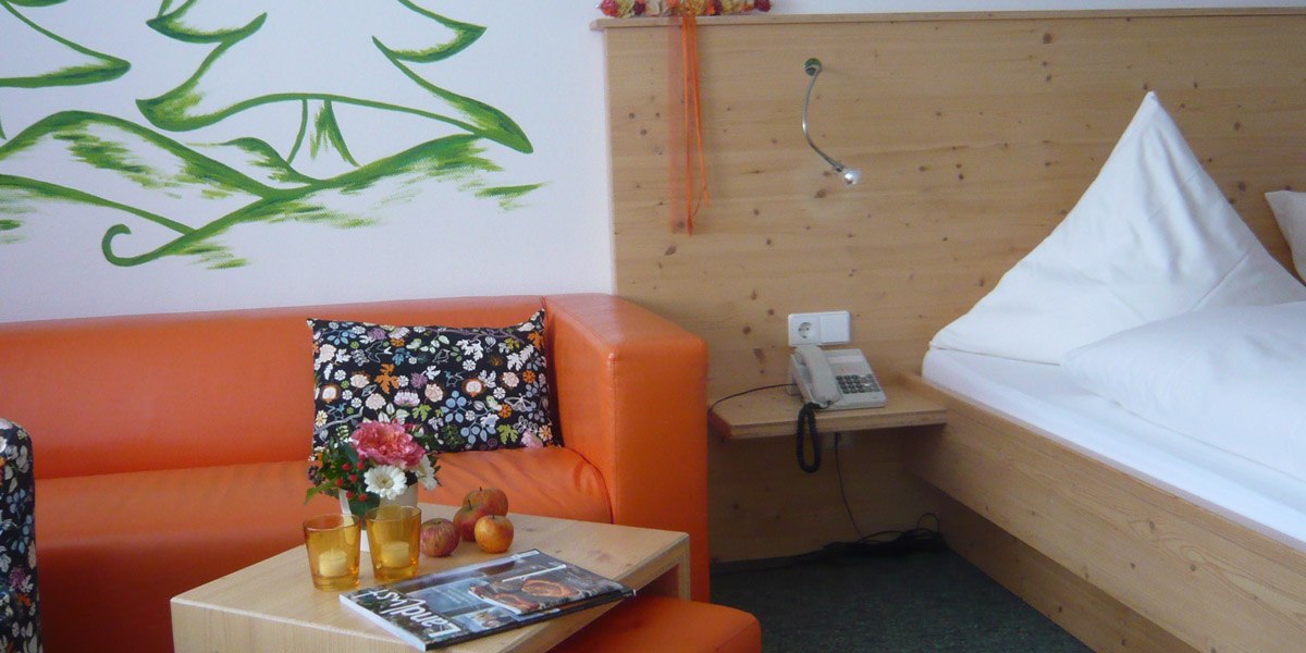 Hotel room with orange sofa, wooden bed, flowers on table, magazine, telephone and green wall painting., &copy; Hotel Hochwald