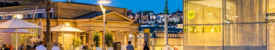 Abendstimmung am Kleinen Schlossplatz mit Palmen, Menschen und moderner Architektur. Im Hintergrund sind beleuchtete Gebäude und eine Statue zu sehen., © SMG, Werner Dieterich
