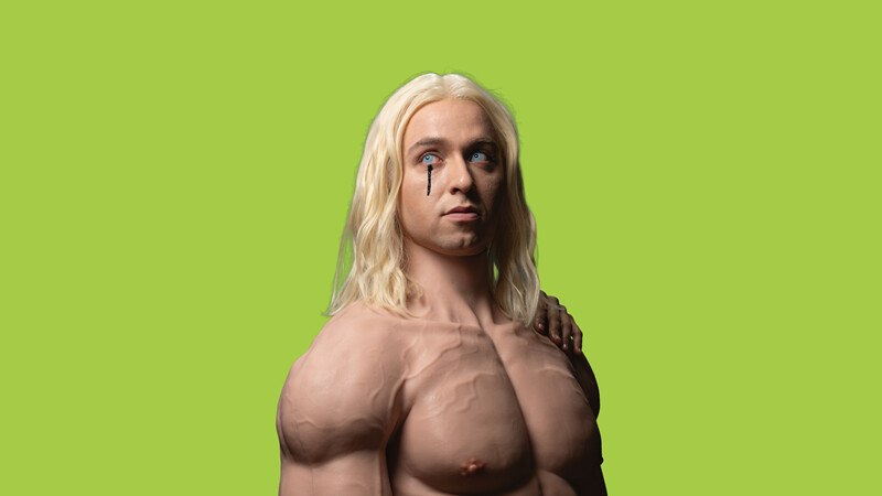 Ein muskulöser Mann mit langen blonden Haaren und einem schwarzen Tränentropfen unter dem Auge vor einem grünen Hintergrund., © Württembergische Staatstheater Stuttgart Ein muskulöser Mann mit langen blonden Haaren und einem schwarzen Tränentropfen unter dem Auge vor einem grünen Hintergrund., © Württembergische Staatstheater Stuttgart