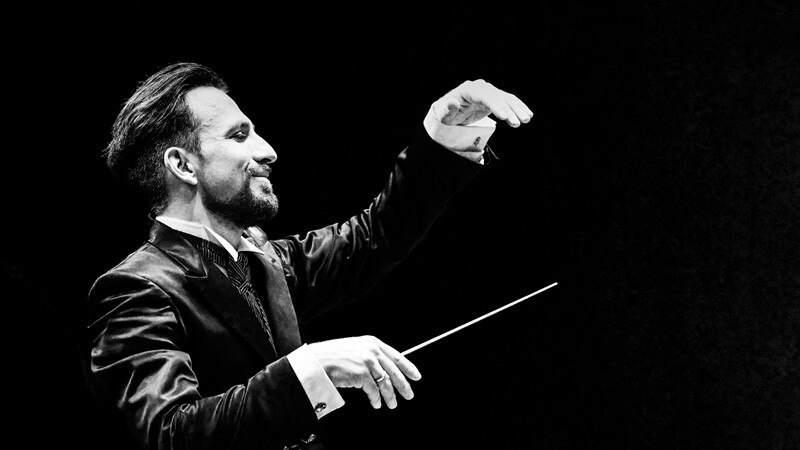 Ein Dirigent in einem eleganten Anzug dirigiert mit einem Taktstock vor einem dunklen Hintergrund. Die Szene ist in Schwarz-Weiß gehalten., © Württembergische Staatstheater Stuttgart Ein Dirigent in einem eleganten Anzug dirigiert mit einem Taktstock vor einem dunklen Hintergrund. Die Szene ist in Schwarz-Weiß gehalten., © Württembergische Staatstheater Stuttgart