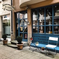 Entrance to a doll's clinic with blue door and shop window. In front of it are potted plants and a white chair. A sign indicates that repairs are being carried out., © Puppen- und Bärenklinik Entrance to a doll's clinic with blue door and shop window. In front of it are potted plants and a white chair. A sign indicates that repairs are being carried out., © Puppen- und Bärenklinik