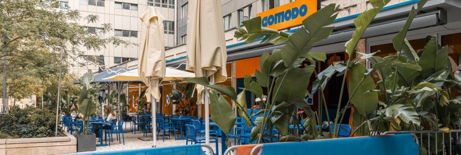 Au&szlig;enbereich eines Caf&eacute;s mit blauen St&uuml;hlen, Tischen und Pflanzen. Das Schild 'comodo' ist sichtbar. Sonnenschirme sind aufgestellt., &copy; Stuttgart Marketing GmbH, Sarah Schmid