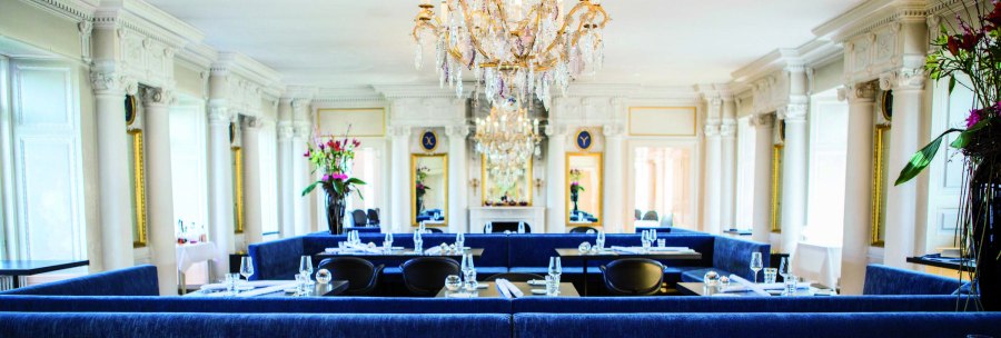 Elegant restaurant with chandelier, blue benches and white pillars. Flower arrangements and laid tables create a luxurious atmosphere., © Speisemeisterei