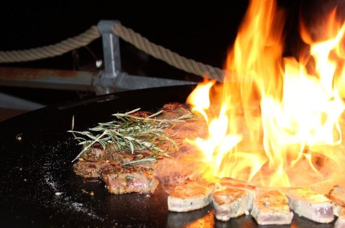 Ein Grill mit brennendem Feuer, Fleischstücken und Rosmarinzweigen im Dunkeln. Im Hintergrund ein Seilgeländer., © Cool-Tours StattReisen Ein Grill mit brennendem Feuer, Fleischstücken und Rosmarinzweigen im Dunkeln. Im Hintergrund ein Seilgeländer., © Cool-Tours StattReisen