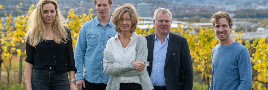 Five people stand smiling in front of an autumnal vineyard. Yellow vine leaves and a cityscape can be seen in the background., © BURKHARDT HELLWIG