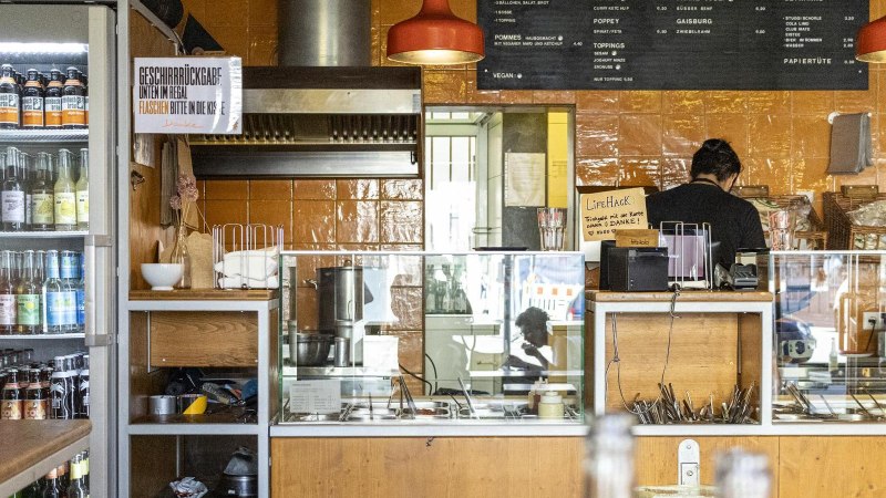 A snack bar with drinks fridge, counter and menu board. One person works behind the counter. The walls are tiled in orange., © SMG, Sarah Schmid