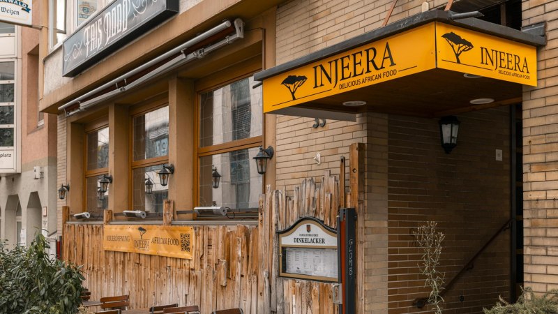 Restaurant Injeera mit gelbem Schild und Holzzaun, bietet afrikanische Küche an. Außenansicht mit Laternen und Pflanzen., © SMG, Sarah Schmid Restaurant Injeera mit gelbem Schild und Holzzaun, bietet afrikanische Küche an. Außenansicht mit Laternen und Pflanzen., © SMG, Sarah Schmid