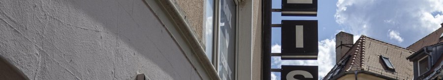 A sign saying 'WHITE' hangs on a building. The sky is blue with a few clouds. Roofs can be seen in the background., © SMG, Sarah Schmid