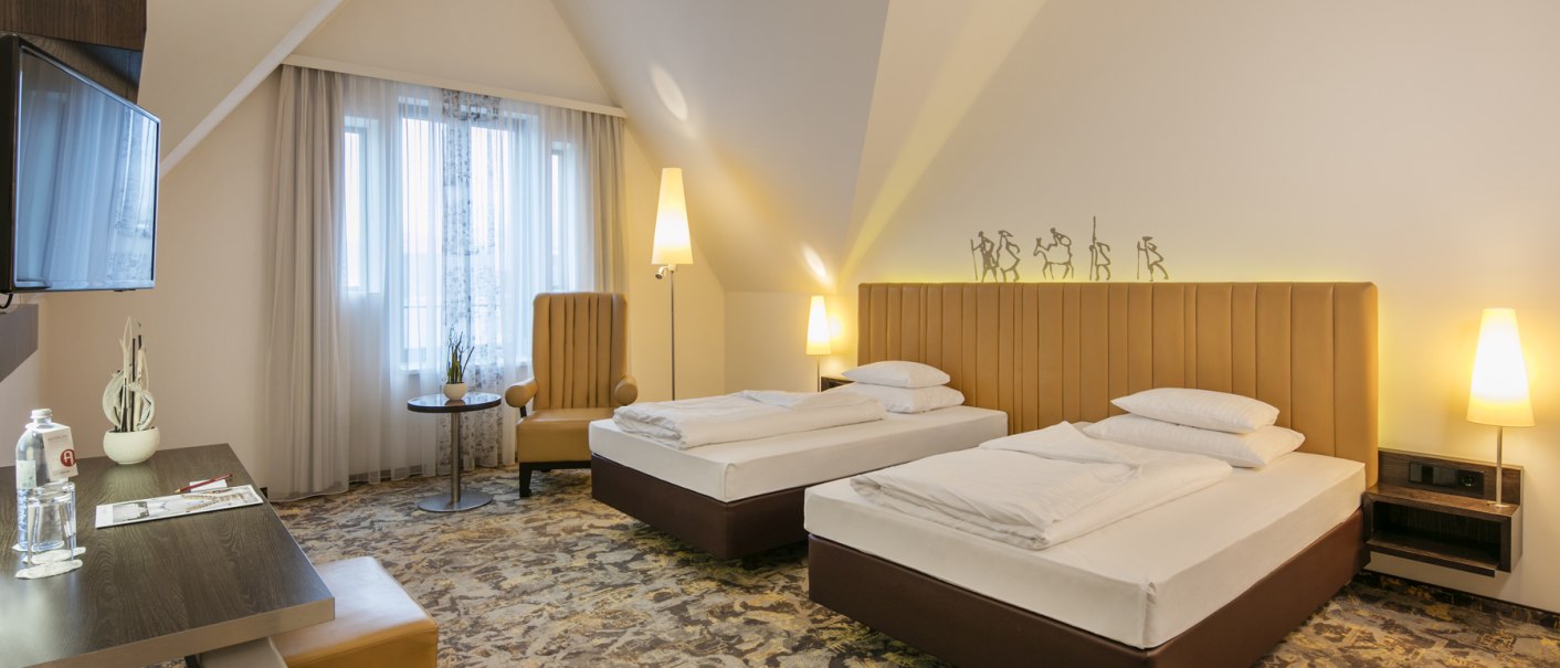 Ein modernes Hotelzimmer mit zwei Einzelbetten, einem Schreibtisch, einem Sessel und einem Flachbildfernseher. Die Einrichtung ist in warmen Tönen gehalten., © ARCOTEL Hotels