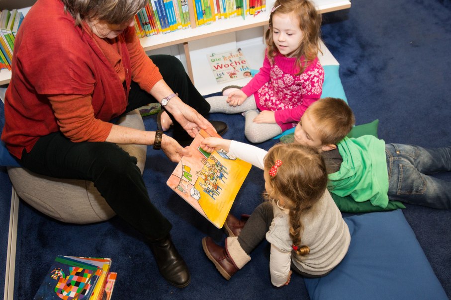 Eine Frau liest drei Kindern in einer Bibliothek ein Bilderbuch vor. Die Kinder sitzen auf Kissen und h&ouml;ren aufmerksam zu., &copy; Stadtbibliothek Stuttgart - Leseohren e.V.