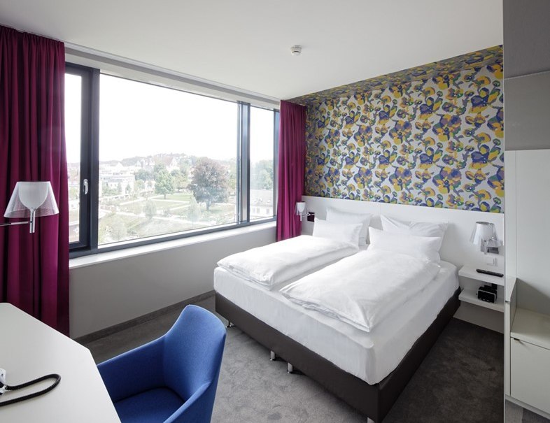 Modern hotel room with double bed, colorful wallpaper, large window and purple curtains. There is a blue chair at the desk., &copy; Hotel am Remspark