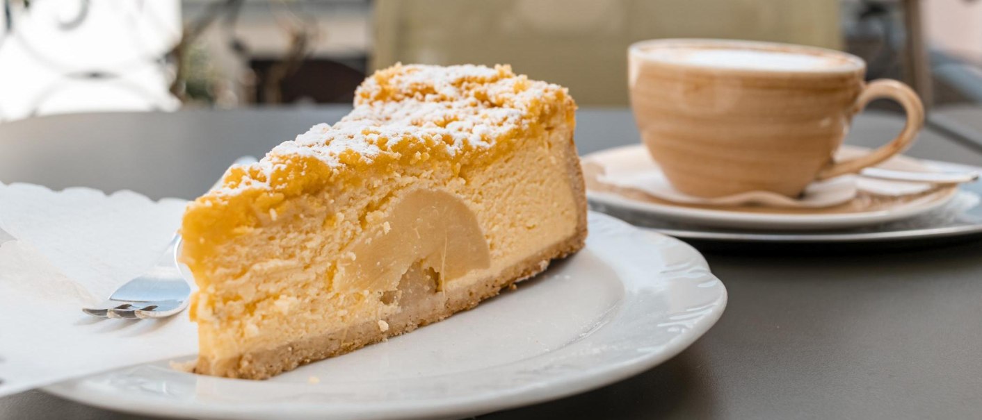 Ein Stück Kuchen mit Puderzucker auf einem Teller, daneben eine Tasse Kaffee auf einem Tisch im Café Schurr., © SMG, Sarah Schmid