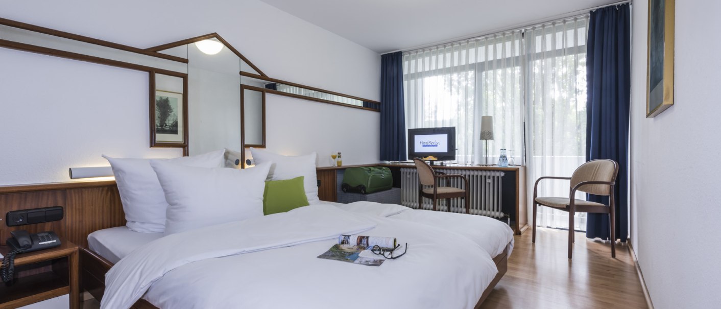A cozy double room with wood accents, a double bed, a desk, a chair, and a large window with curtains. There is a TV on the desk., &copy; Hotel Berlin aZIS Hotel BetriebsGmbH