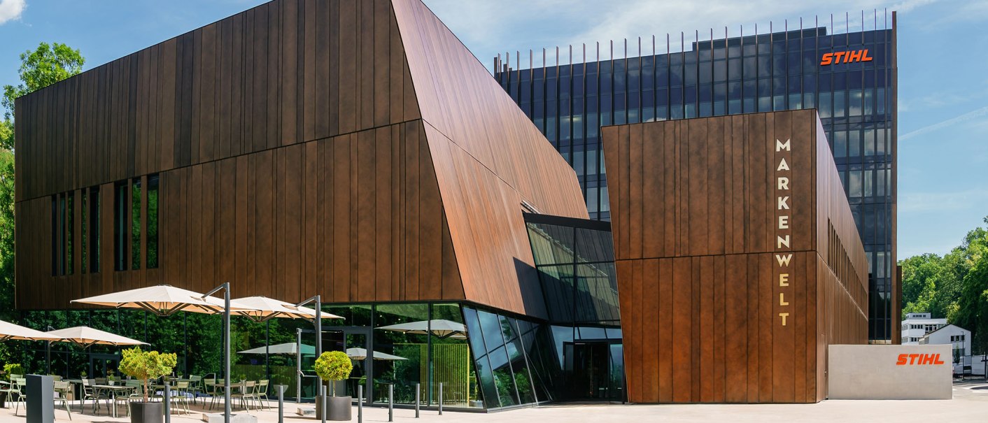 Modern STIHL brand world building with wooden cladding and glass fronts, surrounded by trees. Sunshades and seating in the foreground., © STIHL Markenwelt Modern STIHL brand world building with wooden cladding and glass fronts, surrounded by trees. Sunshades and seating in the foreground., © STIHL Markenwelt