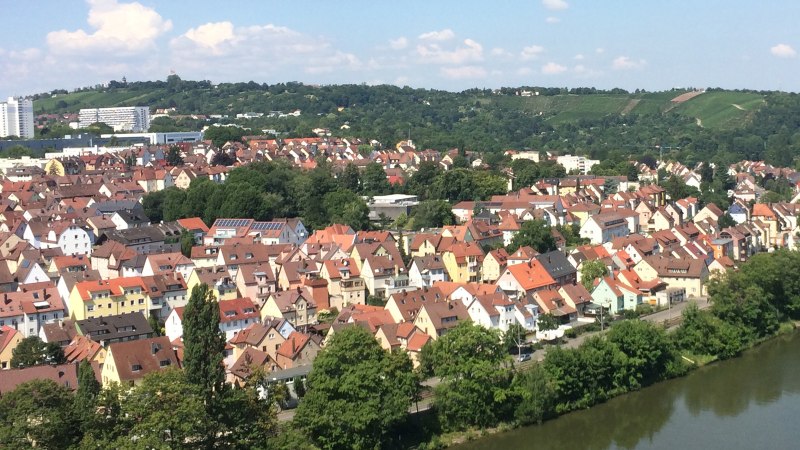 Luftaufnahme einer Stadt mit roten D&auml;chern, einem Fluss und gr&uuml;nen H&uuml;geln im Hintergrund. Der Himmel ist blau mit einigen Wolken., &copy; Aussicht auf den Neckar und die Weinberge