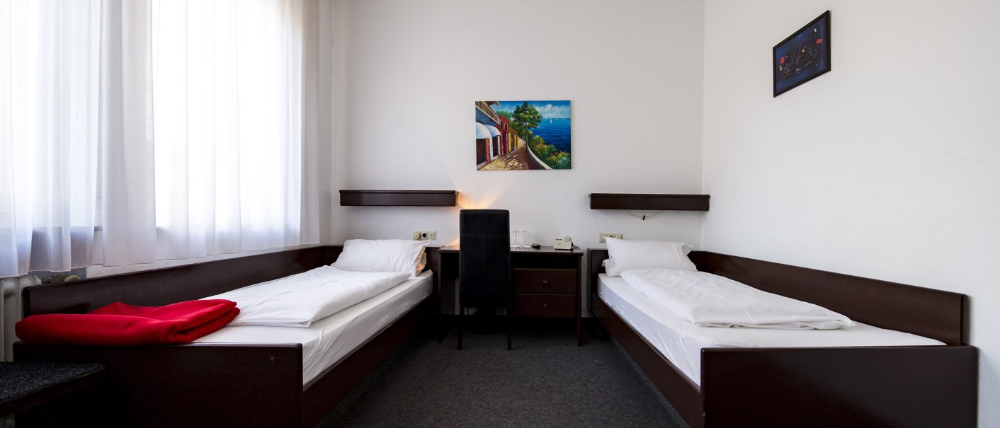 Ein schlichtes Hotelzimmer mit zwei Einzelbetten, einem Nachttisch in der Mitte und zwei Gemälden an der Wand. Ein Fenster mit weißen Vorhängen., © Hotel Ri