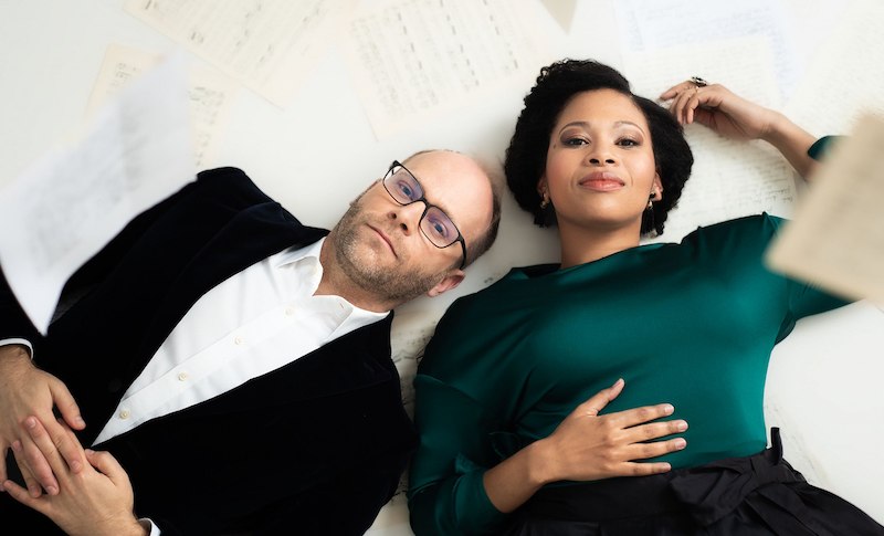 Zwei Personen liegen auf einem wei&szlig;en Hintergrund, umgeben von verstreuten Notenbl&auml;ttern. Sie blicken entspannt in die Kamera., &copy; Internationale Hugo-Wolf-Akademie