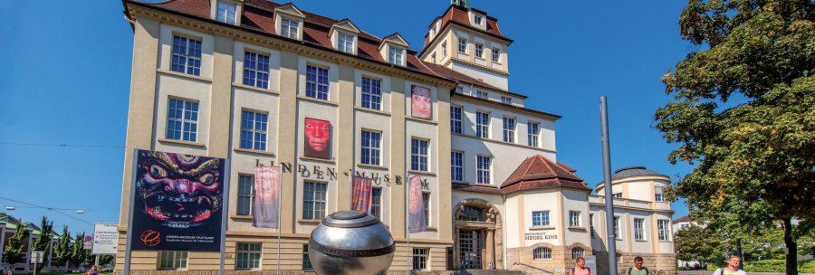 The Linden Museum in Stuttgart in sunny weather. Posters and a large silver sphere can be seen in front of the building. People stroll past., © © Stuttgart-Marketing GmbH