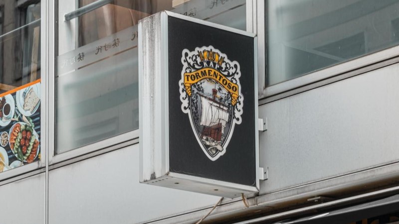 Ein Schild mit dem Logo von Kap Tormentoso hängt an einem modernen Gebäude. Es zeigt ein Segelschiff und dekorative Elemente., © Stuttgart Marketing GmbH, Sarah Schmid