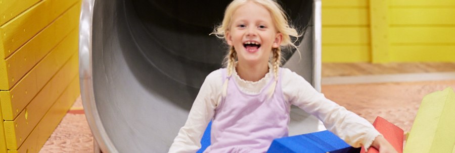 A laughing child slides out of an orange tube on a children's construction site. Surrounded by colorful foam blocks in yellow, blue and red., &copy; Julia Ochs