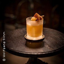 Ein Whiskey Sour in einem Glas auf einem dunklen Holztisch, dekoriert mit einer Waffel und einer Orangenscheibe., © links im Bild