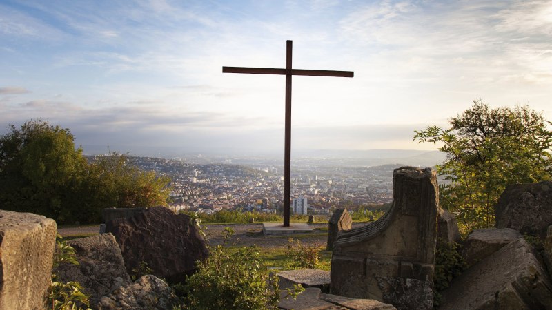 A cross on the Birkenkopf in Stuttgart, surrounded by rubble, with a view of the city in the background at sunset., © Stuttgart-Marketing GmbH, Jean-Claude Winkler A cross on the Birkenkopf in Stuttgart, surrounded by rubble, with a view of the city in the background at sunset., © Stuttgart-Marketing GmbH, Jean-Claude Winkler