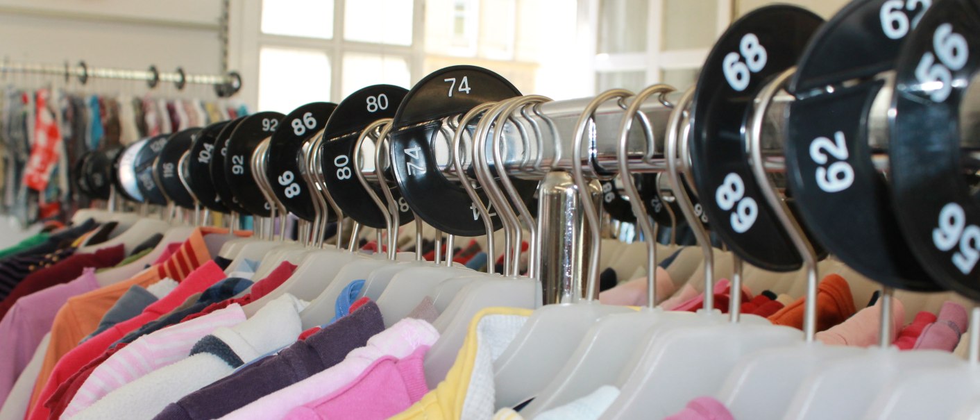 Kleiderstange mit bunter Kinderkleidung und Gr&ouml;&szlig;enmarkierungen in einem Gesch&auml;ft. Die Kleidung ist nach Gr&ouml;&szlig;en sortiert., &copy; ZORA gGmbH