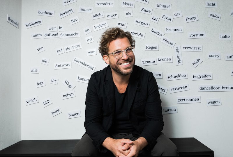 Ein Mann mit Brille sitzt lächelnd vor einer Wand, die mit verschiedenen deutschen Wörtern beklebt ist., © Internationale Hugo-Wolf-Akademie Ein Mann mit Brille sitzt lächelnd vor einer Wand, die mit verschiedenen deutschen Wörtern beklebt ist., © Internationale Hugo-Wolf-Akademie