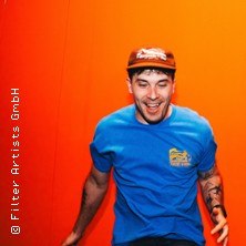 A man in a blue T-shirt and brown cap smiles in front of an orange background., © see on the left A man in a blue T-shirt and brown cap smiles in front of an orange background., © see on the left