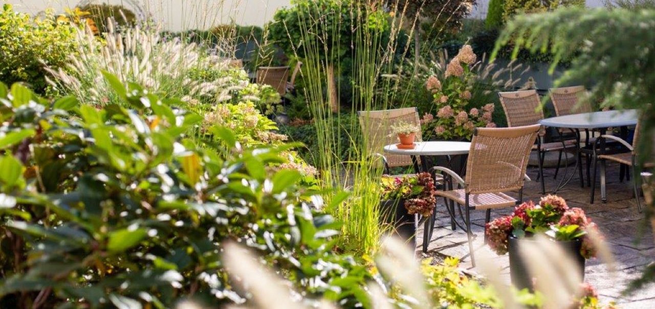 Ein sonniger Garten mit Tischen und Stühlen, umgeben von üppigen Pflanzen und Blumen. Im Hintergrund sind Gebäude zu sehen., © Kronen Hotel GmbH Ein sonniger Garten mit Tischen und Stühlen, umgeben von üppigen Pflanzen und Blumen. Im Hintergrund sind Gebäude zu sehen., © Kronen Hotel GmbH