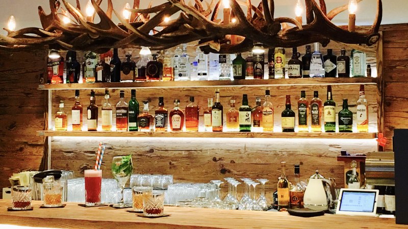 Eine Bar mit einer Holztheke, auf der verschiedene Getränke stehen. Im Hintergrund sind Regale mit Spirituosen und eine Geweih-Lampe zu sehen., © TOMAS Eine Bar mit einer Holztheke, auf der verschiedene Getränke stehen. Im Hintergrund sind Regale mit Spirituosen und eine Geweih-Lampe zu sehen., © TOMAS