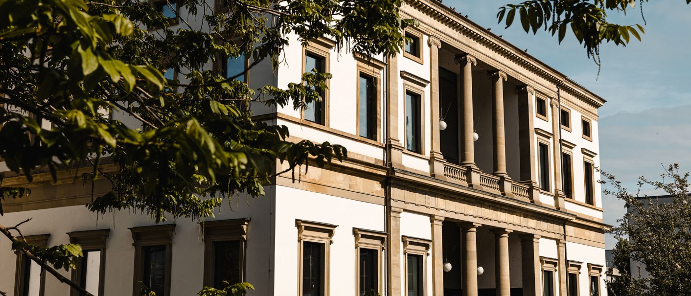 Ein klassisches Gebäude mit Säulen, umgeben von grünen Bäumen. Menschen gehen auf einem Weg vor dem Gebäude entlang., © Stuttgart Marketing GmbH, Sarah Schmid