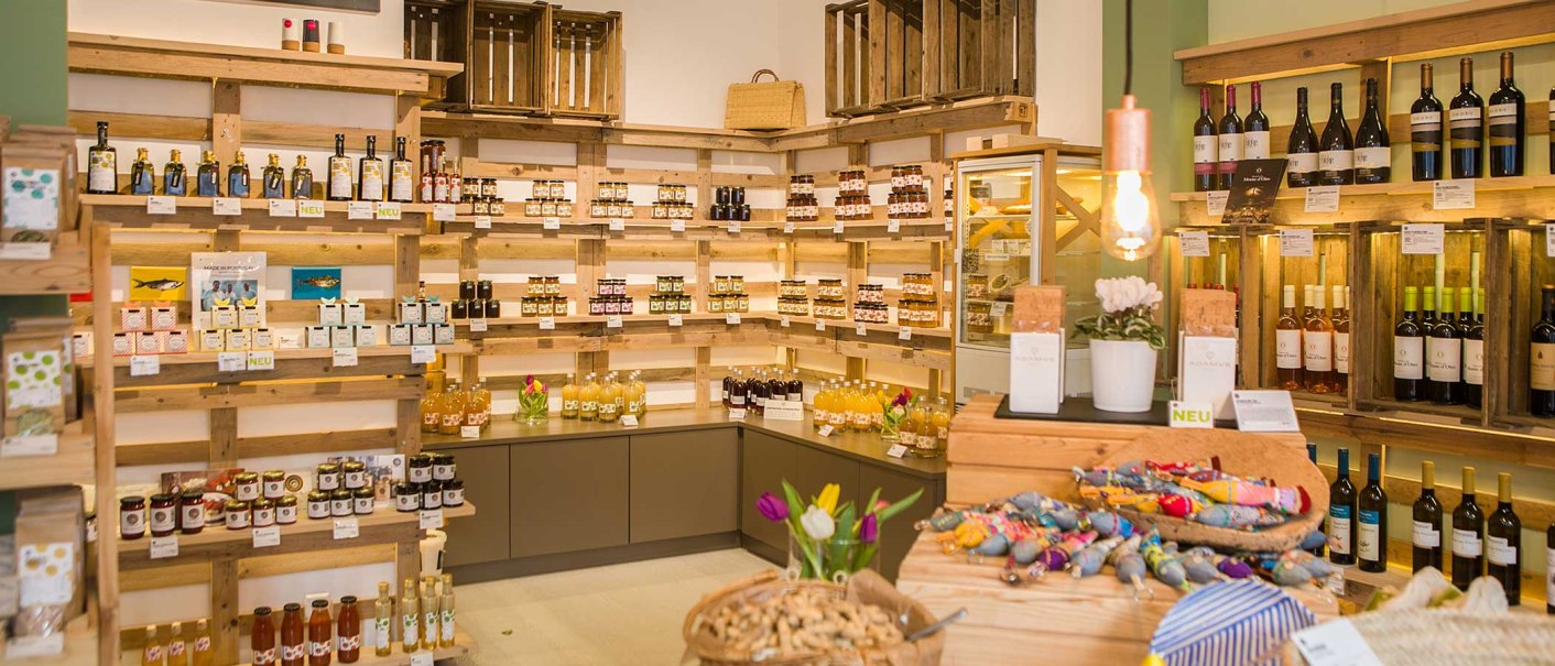 Feinkostladen mit Regalen voller Weinflaschen, Marmeladen und anderen Produkten. Holzkisten und dekorative Elemente schaffen eine gemütliche Atmosphäre., © Benjamin Stollenberg