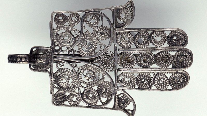 Ein silbernes Hamsa-Amulett mit filigranen, spiralförmigen Mustern. Es symbolisiert Schutz und Glück., © Linden-Museum Stuttgart Ein silbernes Hamsa-Amulett mit filigranen, spiralförmigen Mustern. Es symbolisiert Schutz und Glück., © Linden-Museum Stuttgart