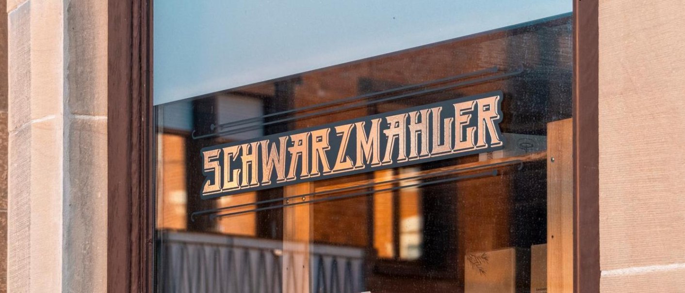 A shop window with the lettering 'Schwarzmahler' in stylized script. The surroundings are reflected in the glass., © SMG Stuttgart Marketing GmbH - Sarah Schmid A shop window with the lettering 'Schwarzmahler' in stylized script. The surroundings are reflected in the glass., © SMG Stuttgart Marketing GmbH - Sarah Schmid