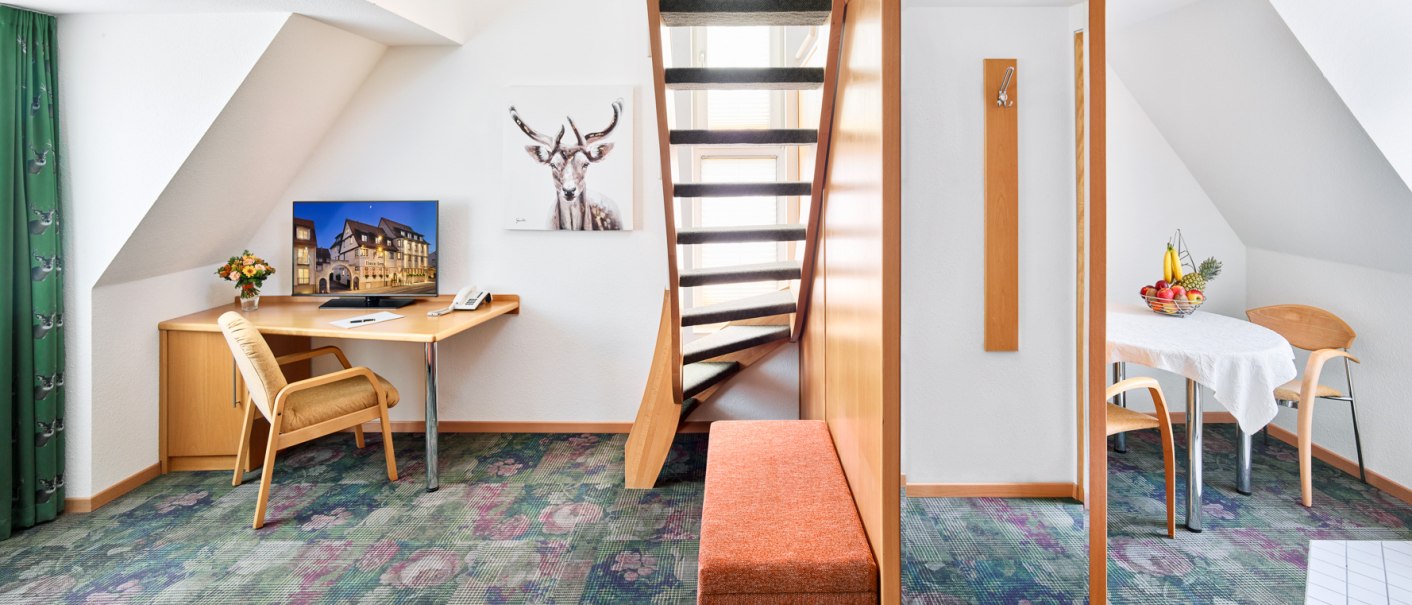 Helles Hotelzimmer mit Schreibtisch, Fernseher, Treppe, Esstisch und Obstkorb. Teppich mit Blumenmuster, Bild eines Hirsches an der Wand., © Hirsch Hotel