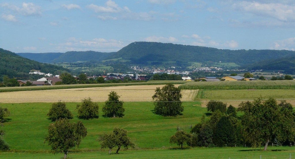 Grüne Felder und Bäume vor einer hügeligen Landschaft am Fuß der schwäbischen Alb, mit einem Dorf im Hintergrund., © Ulli Kellenbenz