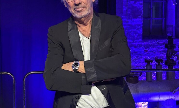 Ein Mann mit Brille und Bart steht lässig mit verschränkten Armen vor einer blauen, industriellen Kulisse., © Renitenztheater Stuttgart e.V. Ein Mann mit Brille und Bart steht lässig mit verschränkten Armen vor einer blauen, industriellen Kulisse., © Renitenztheater Stuttgart e.V.