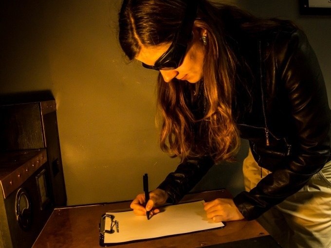 Woman with sunglasses writing on a clipboard in a dimly lit room. She is wearing a black jacket., © ExitGames Stuttgart Woman with sunglasses writing on a clipboard in a dimly lit room. She is wearing a black jacket., © ExitGames Stuttgart