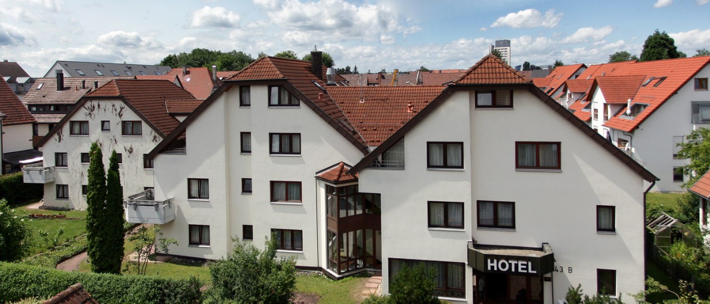 A hotel with a white façade and red roofs, surrounded by gardens and other houses under a cloudy sky., © Hotel Flora A hotel with a white façade and red roofs, surrounded by gardens and other houses under a cloudy sky., © Hotel Flora