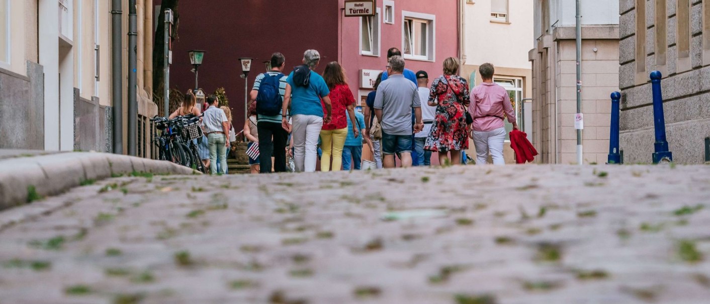 Menschen gehen auf einer gepflasterten Straße im Bohnenviertel. Im Hintergrund sind Gebäude und ein Schild mit der Aufschrift 'Hotel Türmle' zu sehen., © Thomas Niedermüller Menschen gehen auf einer gepflasterten Straße im Bohnenviertel. Im Hintergrund sind Gebäude und ein Schild mit der Aufschrift 'Hotel Türmle' zu sehen., © Thomas Niedermüller